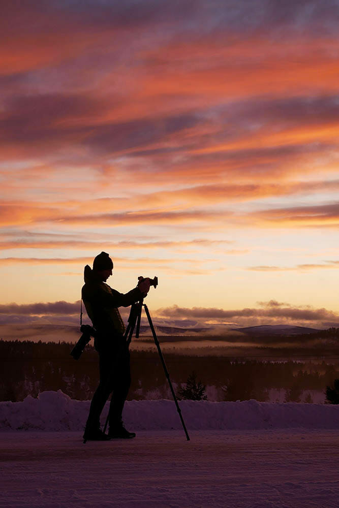 A silhouette of a young photographer shooting a snowy landscape at sunset with a camera on a tripod